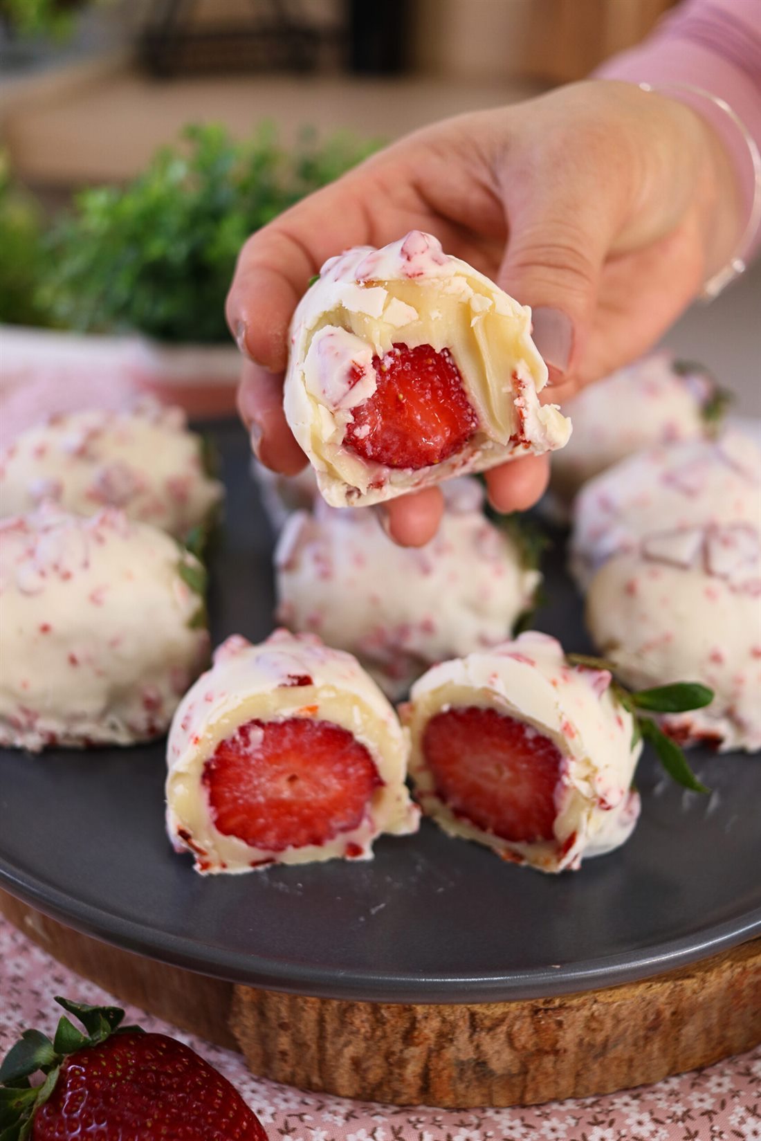 Mão segurando um morango cravejado partido ao meio: a fruta vermelha no centro, envolvida por brigadeiro branco e casca de chocolate branco cravejada de lascas rosadas. Ao fundo, outras unidades sobre um prato escuro.